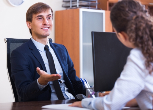 A man in a suit is sitting at a desk, gesturing while discussing financial matters with a person across the table. 