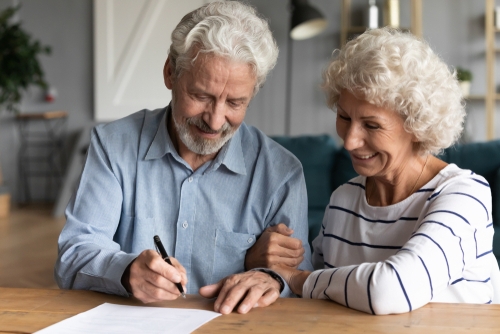 An elderly man is thoughtfully signing a will-based estate plan at the table, while an elderly woman looks on with a warm smile.