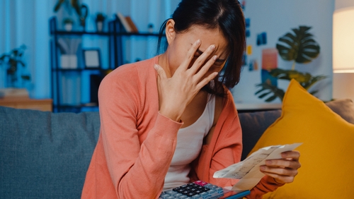 A person sits on a couch, overwhelmed, covering their face with a receipt in hand. Beside them, a calculator hints at financial worries. In the background, shelves and a yellow pillow offer little comfort to the lingering concern.