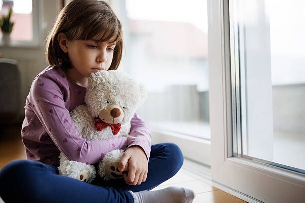 Sad lonely little girl hugging her teddy bear by the window