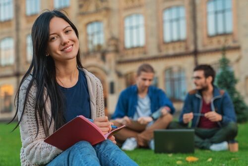 Portrait of young female student woman girl noting doing homework task sitting on the grass while two males college students classmates boys using laptop in background at university campus lawn