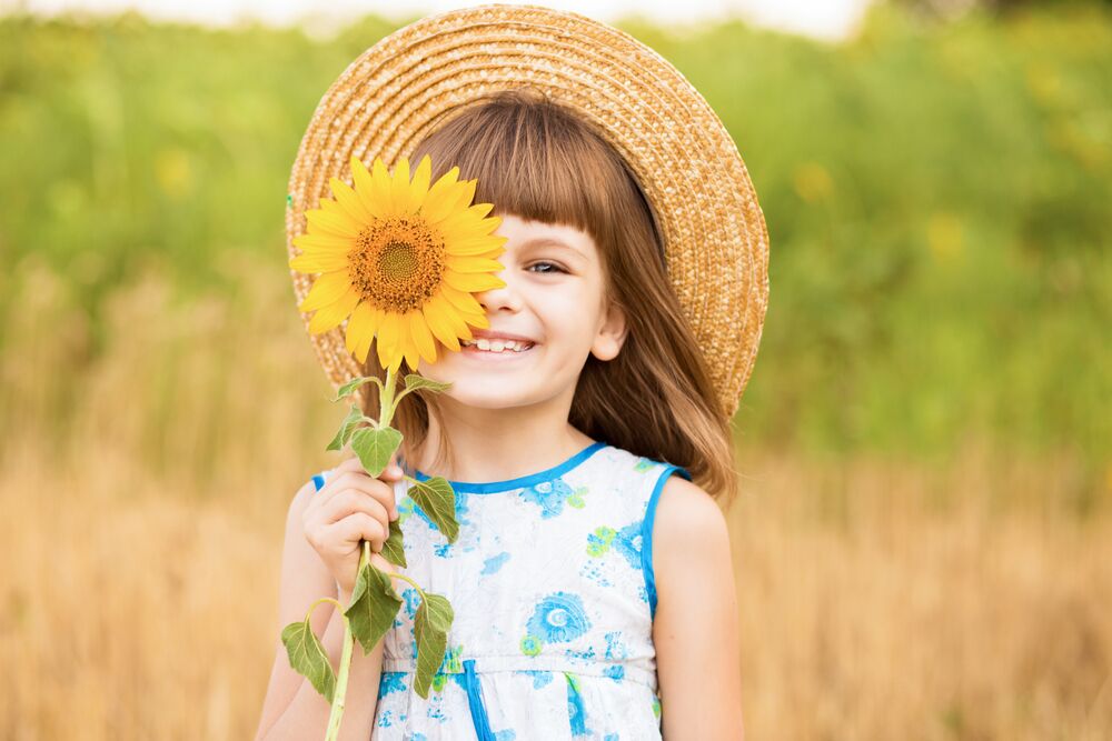 Beautiful little girl in straw hat with fluttering hair smile and hide eye with sunflower flower, walking outdoor in summer holiday