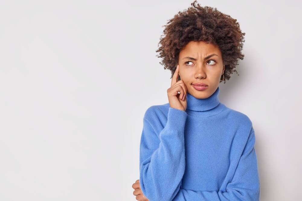 A women standing in front of a grey background with her finger scratching the side of her head and her eyes are looking to the side