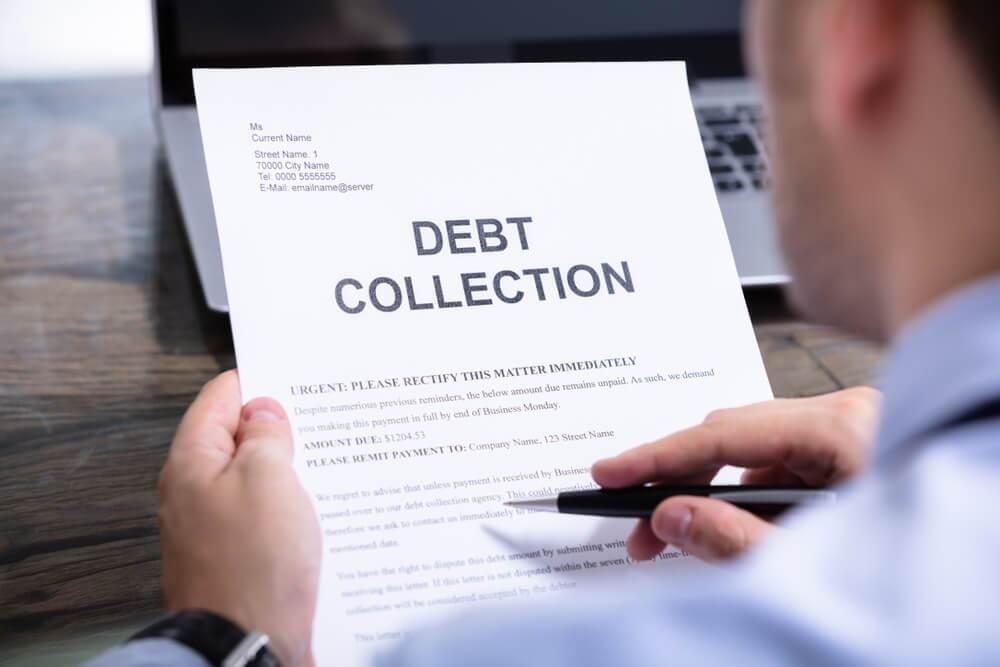 A man sitting at a desk reading a paper that says debt collections with a pen in his right hand
