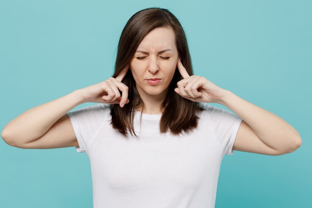 A woman standing against a blue background with her fingers covering her ears and her eyes closed