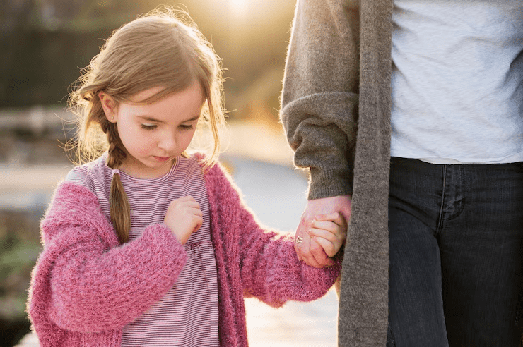 A young girl in a pink sweater holds an adult's hand while looking down, standing outdoors in sunlight, while her parent wonders how is child support calculated in Texas.