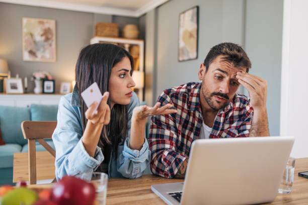 An adult cheerful couple sitting at the table, using a laptop, with the living room visible in the background. They are shopping online with a credit card and look concerned.