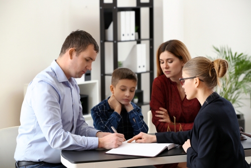 In an office setting, amidst the tension of a custody battle, a man cautiously signs a document at the table, observed by a woman, while another woman points to key sections. A child stands nearby, highlighting what should you not do during such crucial moments.