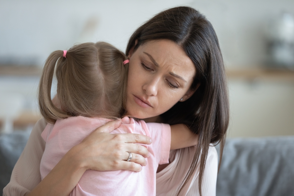 A woman with a concerned expression hugs a young girl with pigtails, both wearing light pink shirts, sitting indoors