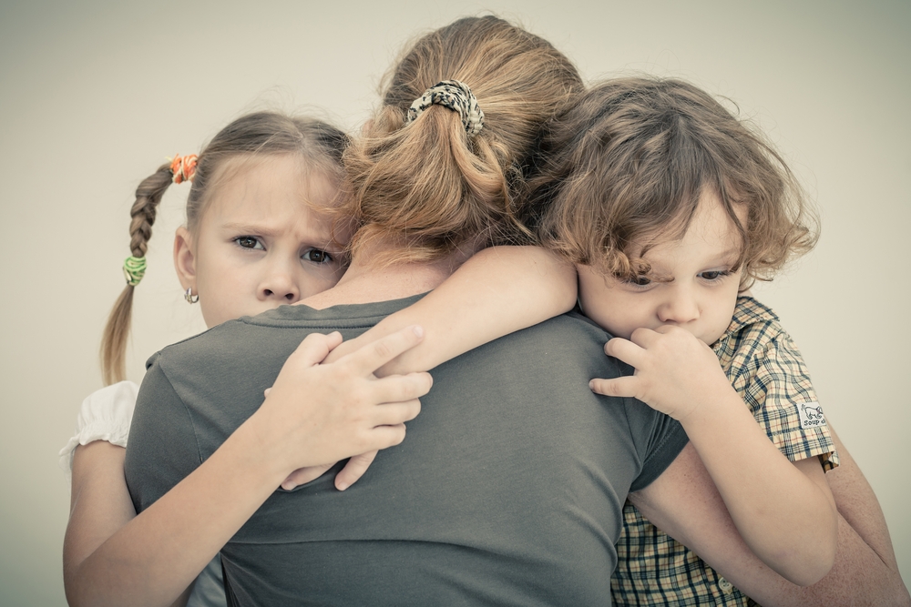 A woman seen from behind hugs two young children, a girl and a boy, who appear anxious and are clinging to her