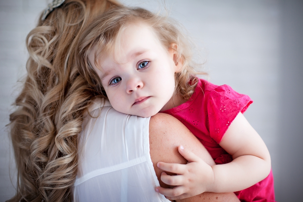A young girl in a pink dress rests her head on a woman's shoulder, looking at the camera while being held
