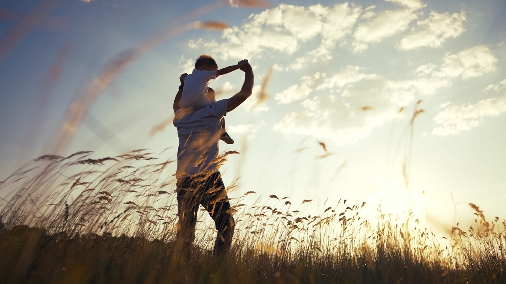 An adult, perhaps a custodial parent not following court order in Texas, carries a child on their shoulders while walking through a grassy field at sunset beneath a partly cloudy sky.