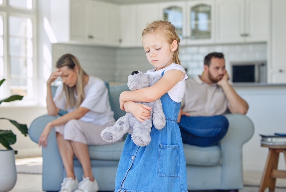 A young girl hugs a stuffed animal while two adults sit apart on a couch in the background, both appearing upset, raising questions like how can a mother lose custody in Texas.