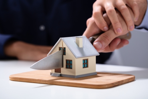 A hand holding a knife slices through a small model house placed on a wooden board, symbolizing the division of property during the forced sale of a house in divorce.