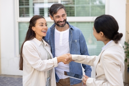 A couple shakes hands with a woman holding a clipboard, standing outside a building with green window blinds, as they discuss the logistics of forcing the sale of their house in divorce proceedings.