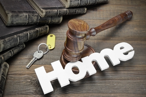 A wooden gavel rests on a table next to a key tagged with "Home," hinting at the complexity of dividing assets. Old books lie stacked nearby, reminiscent of past decisions and the legal intricacies surrounding the forcing sale of a house in divorce.