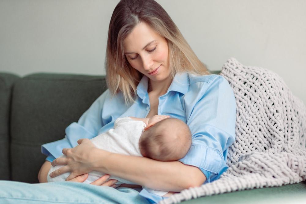 A woman in a blue shirt sits on the couch, breastfeeding her baby, while a knitted blanket drapes behind. She wonders if a father can get full custody of a newborn, reflecting on family possibilities as she nurtures her child in the serene setting.