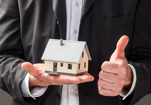 A person in a suit holds a small model of a house in one hand, proudly giving a thumbs-up with the other, as they promote the concept of placing your house in a trust to protect your legacy.