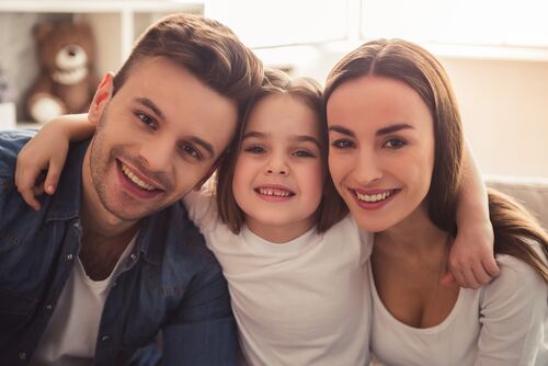 Charming little girl and her beautiful young parents are hugging, looking at camera and smiling while sitting at home