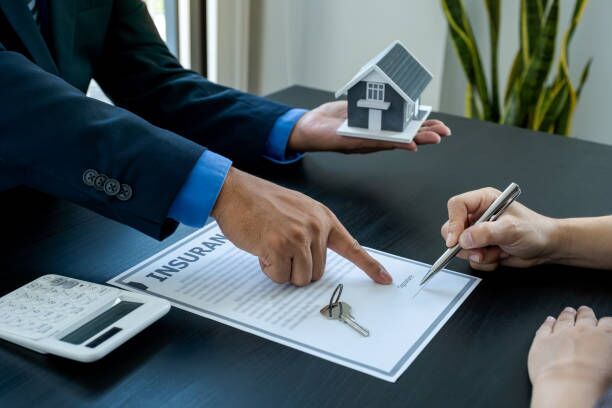 Person signing a home insurance contract in front of an agent pointing where to sign and holding a miniature house in the other hand