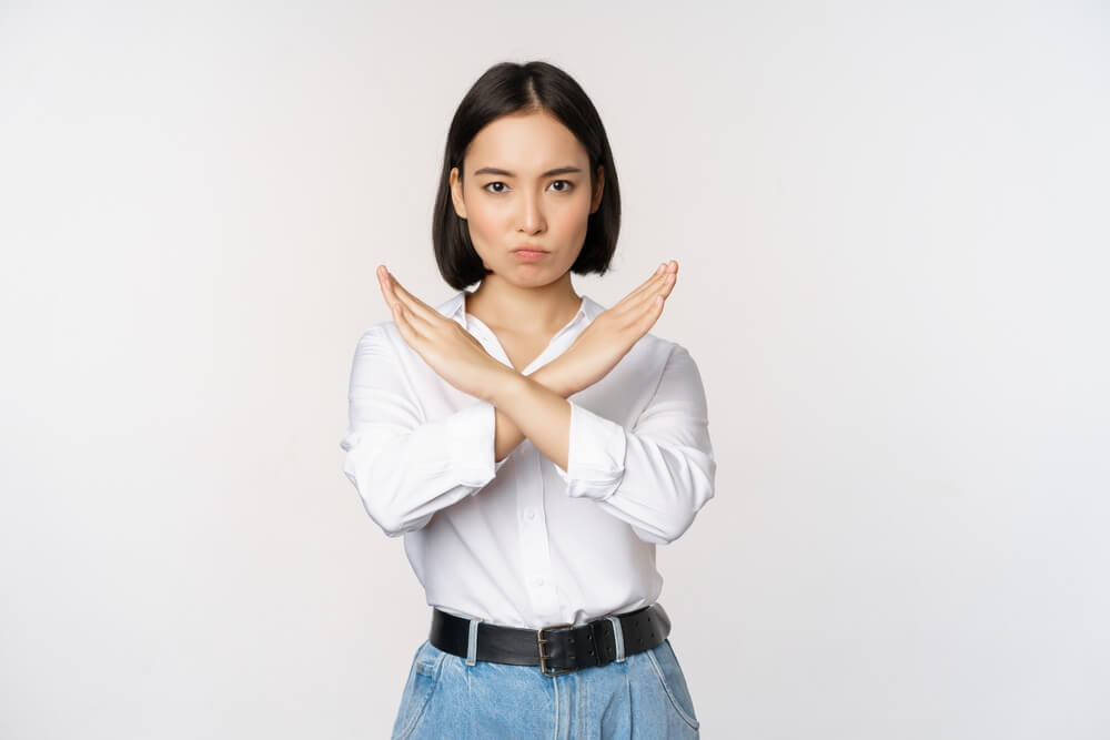 A woman standing against a white wall holding both of her hands up in a X shape showing a sign of refusal