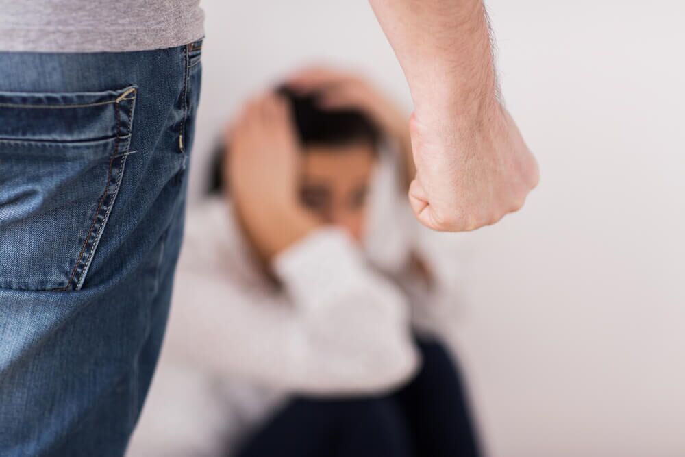 A woman sitting on the ground covering her head because she is scared and there is a man with his hand clinched in a fist