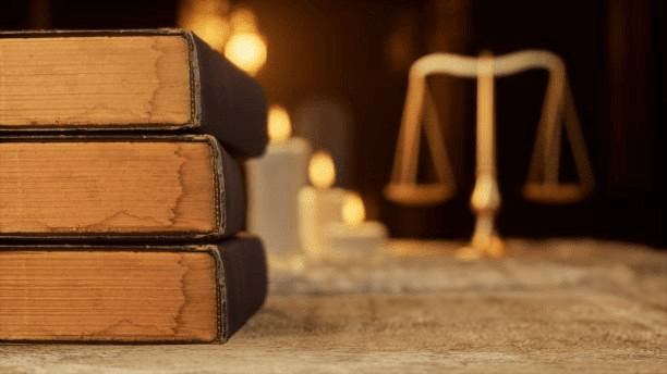 “Stack of vintage legal books on a desk with a blurred background featuring scales of justice and warm candlelight, symbolizing law and justice.”
