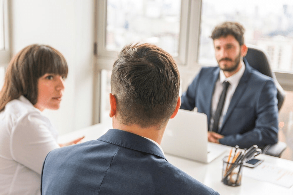 Three people in business attire are having a meeting in a bright office, with a laptop and documents on the table. Two are facing each other while one, with their back to the camera, is listening intently. 