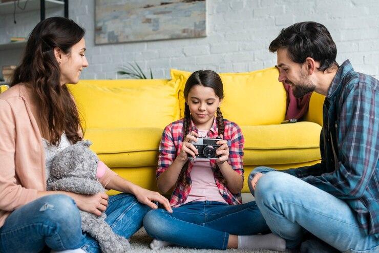 A young girl shows a camera to her parents in a living room, all sitting on the floor, with a yellow couch and a fluffy toy nearby, after temporary custody was granted.